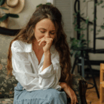 Woman sitting on a couch with her hand covering her mouth, appearing emotional and distressed in a quiet living room.