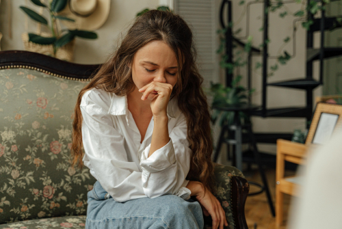 Woman sitting on a couch with her hand covering her mouth, appearing emotional and distressed in a quiet living room.