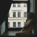 A woman sitting alone by a window, looking out thoughtfully, reflecting on the question of how can I afford to live alone after divorce.