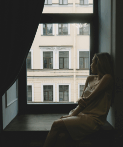 A woman sitting alone by a window, looking out thoughtfully, reflecting on the question of how can I afford to live alone after divorce.
