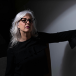 Confident middle-aged woman with silver hair and glasses, standing in dramatic light with her hand against a wall, conveying strength and reflection.