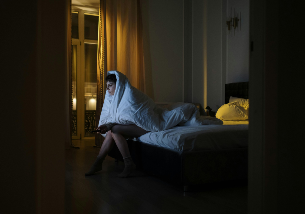 Woman sitting alone on the edge of a bed at night, wrapped in a blanket and looking out a window, appearing anxious and deep in thought.