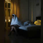 Woman sitting alone on the edge of a bed at night, wrapped in a blanket and looking out a window, appearing anxious and deep in thought.