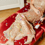 Woman in an elegant gold evening gown reclines on a red chaise lounge with confetti scattered around her, holding a champagne bottle, suggesting celebration or aftermath of a party.