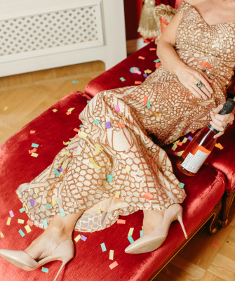 Woman in an elegant gold evening gown reclines on a red chaise lounge with confetti scattered around her, holding a champagne bottle, suggesting celebration or aftermath of a party.