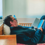 Man lying on a bed reading a book, representing comfort and staying in a familiar but unhappy marriage