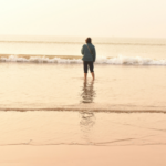 Woman standing alone in shallow ocean water at the beach, looking out toward the waves at sunrise or sunset.
