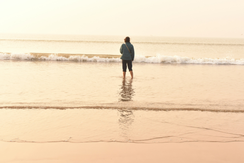 Woman standing alone in shallow ocean water at the beach, looking out toward the waves at sunrise or sunset.